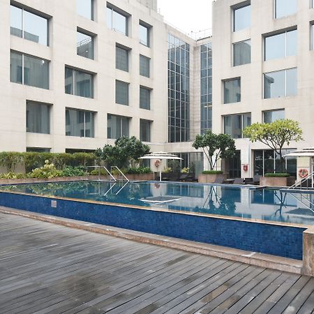 Holiday Inn New Delhi Aerocity By Ihg Ngoại thất bức ảnh The photo shows a modern outdoor swimming pool situated in a courtyard surrounded by several buildings. The pool has a sleek design with a blue tiled edge and is bordered by wooden decking. There are a few trees and plants around the area, adding som